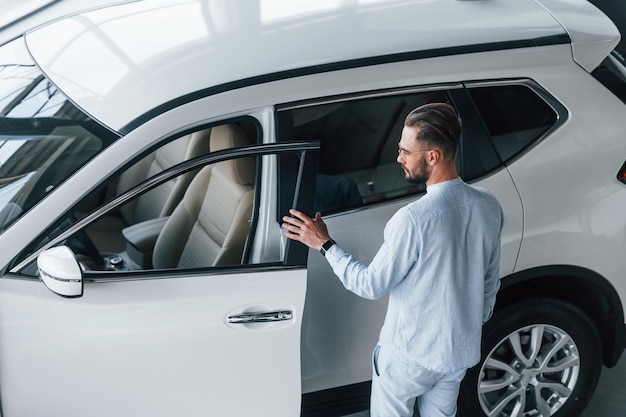 Hombre elegante joven en ropa formal blanca de pie cerca de automóvil blanco