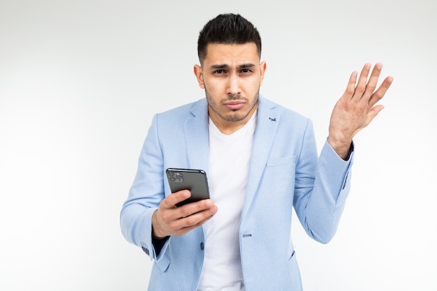 Hombre elegante morena con una chaqueta clásica azul está estudiando sospechosamente información en el teléfono sobre un fondo blanco.