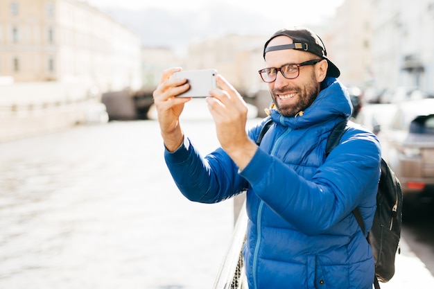 Hombre elegante con ojos azules encantadores y barba con anorak azul, gafas y gorra haciendo selfie con teléfono móvil