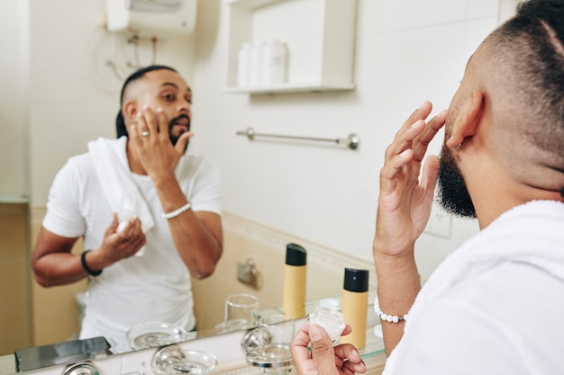 Hombre elegante de pie en el espejo en el baño y aplicar loción humectante en la cara