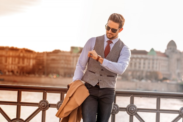 Un hombre elegante de pie junto a un río y mirando la hora