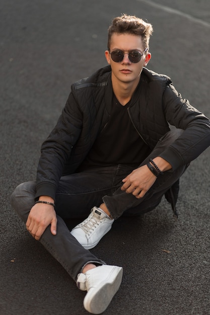 Hombre elegante en ropa de moda negra con gafas de sol en chaqueta de moda negra con zapatillas blancas sentadas en el suelo en la calle