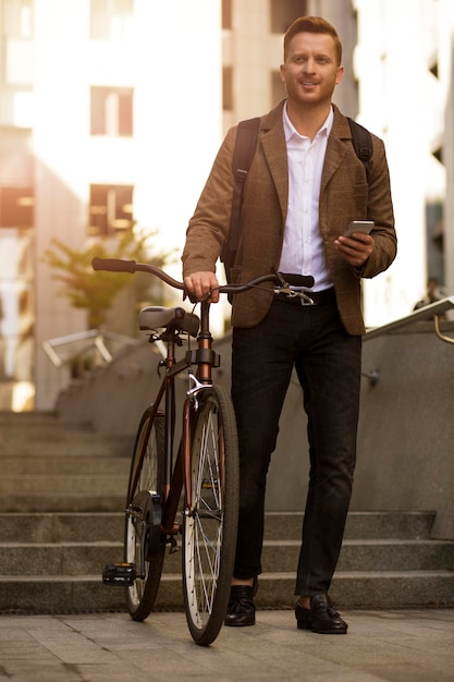 Hombre elegante sonriente caminando su bicicleta con un teléfono móvil en la mano