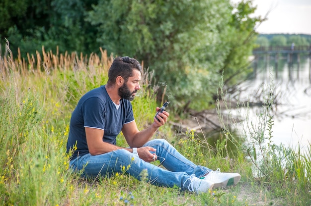 Hombre elegante vape brutal fumando un cigarrillo electrónico en la hierba del bosque