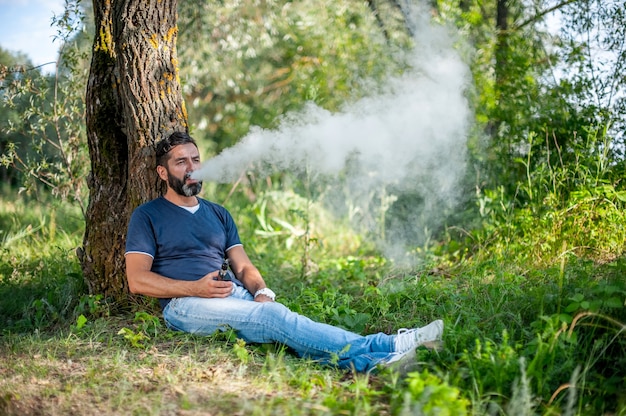 Hombre elegante vape explota un par de cigarrillos electrónicos en el bosque. Hombre guapo.