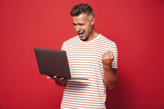 Hombre emocionado de 30 años en camiseta a rayas sonriendo y sosteniendo un portátil gris aislado en rojo