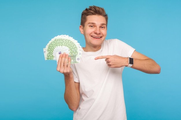 Un hombre emocionado con una camiseta blanca feliz de tener mucho dinero apuntando a los billetes de euro en la mano y sonriendo con entusiasmo disfrutando de un inesperado premio de lotería en un estudio interior aislado en un fondo azul