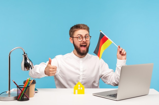 Hombre emocionado positivo con anteojos mostrando los pulgares hacia arriba sosteniendo la bandera de alemania, sentado en el lugar de trabajo con una casa de papel, satisfecho con la compra de alojamiento. Disparo de estudio interior aislado sobre fondo azul.