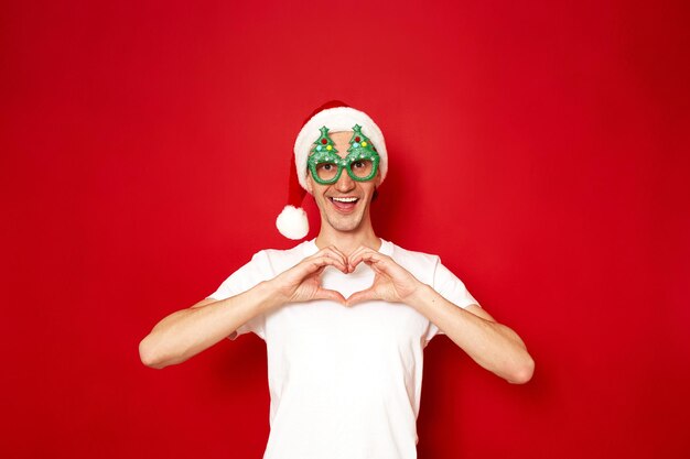 Hombre emocionado con sombrero de año nuevo de Papá Noel y gafas de árbol de Navidad con las manos juntas en forma de corazón