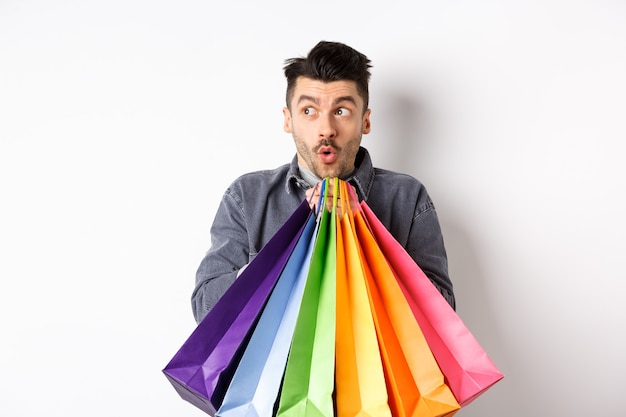 Hombre emocionado sosteniendo coloridas bolsas de la compra y mirando a la izquierda una oferta promocional especial en la tienda, de pie sobre fondo blanco.