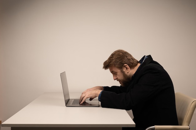 Hombre emocional de barba en posición encorvada sentado en la sala de oficina, trabajando con una computadora portátil