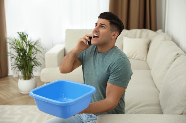 Hombre emocional llamando al servicio de reparación de techos mientras recoge agua del techo en la sala de estar