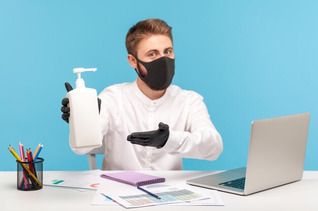 Hombre empleado con mascarilla higiénica y guantes apuntando con botella con gel antiséptico, recomendando desinfección para prevenir el coronavirus en el lugar de trabajo, protección contra enfermedades contagiosas en estudio de oficina