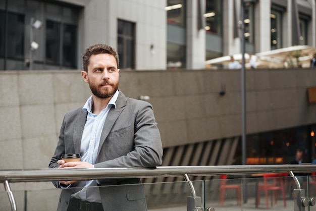 Hombre empresario exitoso en ropa formal mirando a un lado, mientras está de pie y bebiendo café para llevar frente al moderno centro de negocios