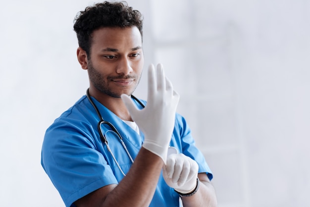 Hombre encantado positivo manteniendo una sonrisa en su rostro mientras usa guantes estériles y está parado sobre blanco