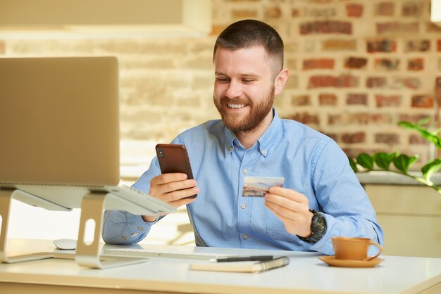 Un hombre encuentra una tienda en línea en un teléfono inteligente con una tarjeta de crédito en la mano frente a la computadora en casa