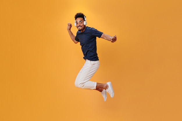 Hombre enérgico en traje elegante saltando sobre fondo naranja y escuchando música en los auriculares Cool guy un blue shirt divirtiéndose en un fondo aislado