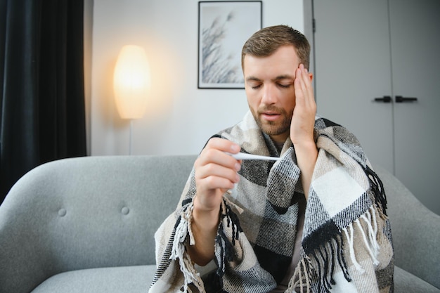 Hombre enfermo acostado en el sofá controlando su temperatura debajo de una manta en casa en la sala de estar
