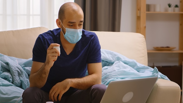 Hombre enfermo en casa hablando en línea con su médico, sosteniendo una botella con pastillas