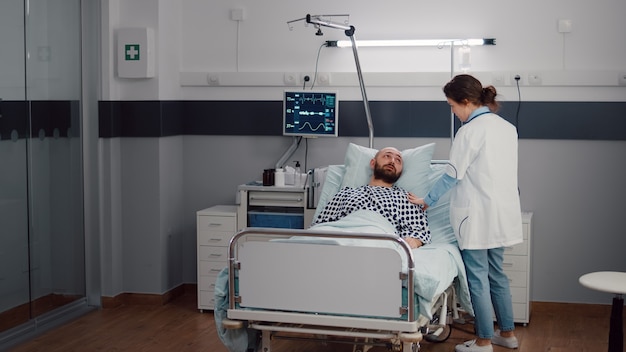 Hombre enfermo descansando en la cama mientras el médico terapeuta monitorea la recuperación respiratoria trabajando en la sala del hospital