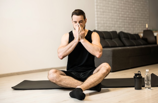 Hombre enfermo con secreción nasal sentado en la estera de yoga negra. Concepto de estancia en casa. Joven morena se suena la nariz después del entrenamiento. Practicar deporte en casa si te sientes mal.