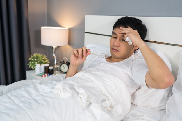 El hombre enfermo tiene dolor de cabeza y usa un termómetro para controlar su temperatura en la cama