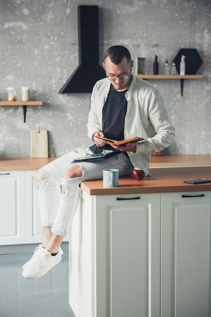 Hombre enfocado usando tecnología moderna trabajando desde casa sentado en la mesa en la cocina buscando información en un libro Freelancer adicto al trabajo estilo de vida exitoso Retrato masculino Estudio de trabajo remoto en casa