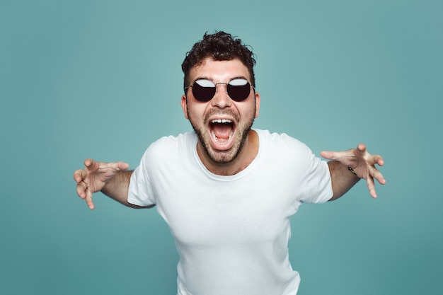 Hombre enojado con gafas de sol gritando y gritando sobre la pared azul