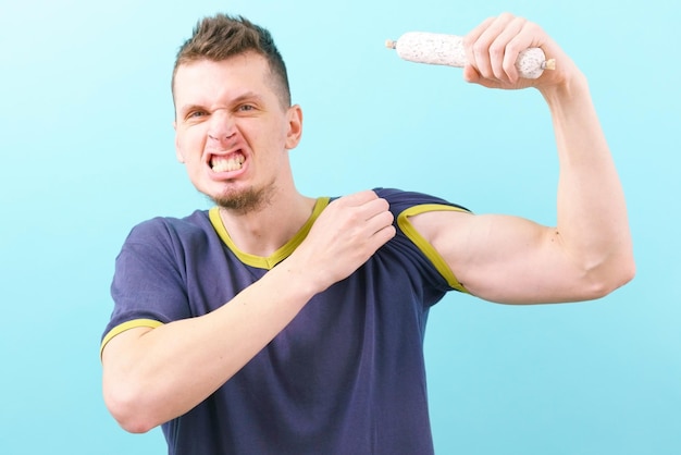 Un hombre enojado que sostiene una salchicha de fuet blanca y muestra sus bíceps en un azul