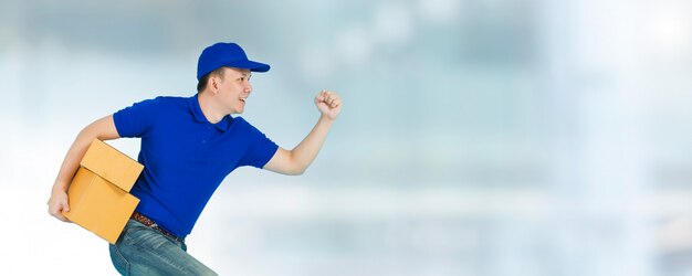 Hombre de entrega feliz asiático vistiendo una camisa azul corriendo y llevando cajas de paquetes de papel aisladas en la ventana de desenfoque.