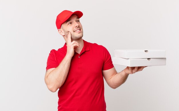 Hombre de entrega de pizza sonriendo felizmente y soñando despierto o dudando, mirando hacia el lado