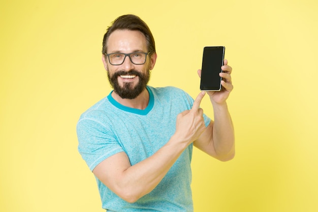 Hombre entusiasmado con las nuevas oportunidades móviles. Chico anteojos alegre apuntando al teléfono inteligente. Aplicación de interacción de usuario feliz de hombre para teléfono inteligente. Guy barbudo interactúa con la aplicación de interfaz móvil.