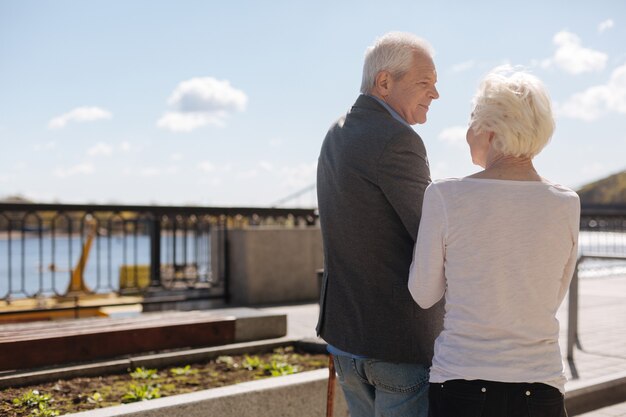 Hombre entusiasta alegre de cabeza gris sonriendo y escuchando a su esposa mientras las mujeres de edad hablando