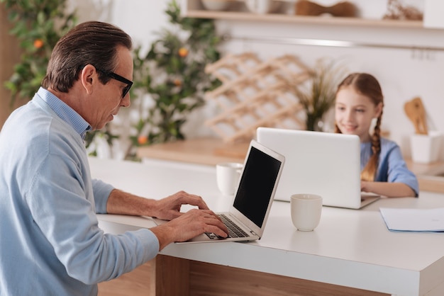 Hombre envejecido agradable jubilado sentado en casa y usando computadoras portátiles con el niño mientras envía mensajes de texto y revisa los perfiles de las redes sociales