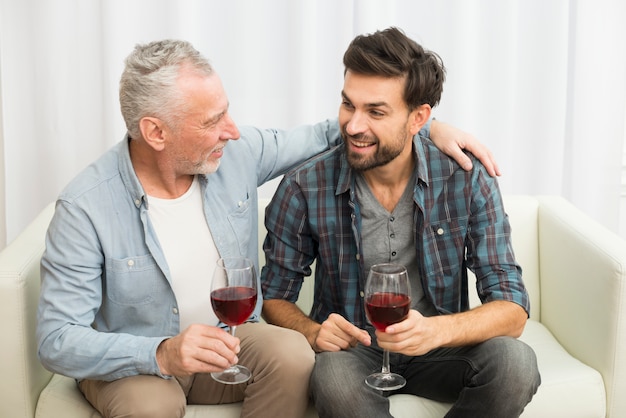 Hombre envejecido que abraza al individuo sonriente joven con los vidrios de vino en el sofá