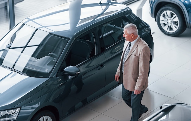 Hombre envejecido en ropa formal caminando en el salón del automóvil y buscando el coche.