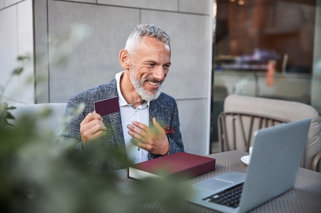 Hombre de envejecimiento enérgico sosteniendo una tarjeta de plástico de color rojo oscuro y gesticulando con la mano delante de la computadora portátil