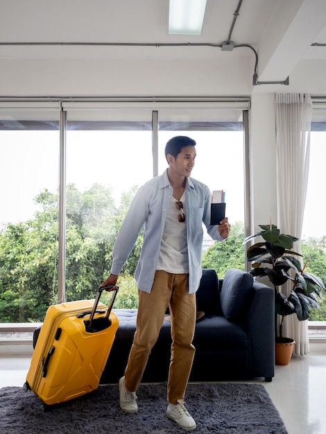 Un hombre con equipaje amarillo Guapo hombre asiático en camisa de mezclilla caminando mientras sostiene el pasaporte cerca de la ventana de vidrio estilo vertical Concepto de vacaciones de viaje de vacaciones de verano Tiempo para viajar