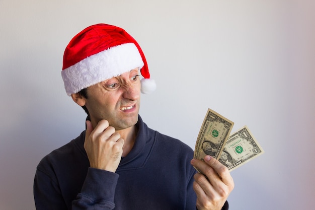 Hombre escéptico con sombrero rojo de Navidad sosteniendo billetes de un dólar con mirada horrorizada