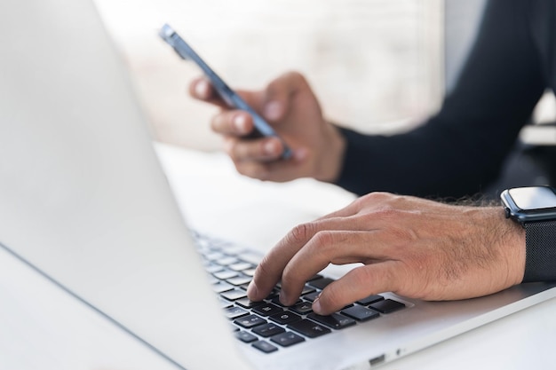 Foto el hombre escribe en el teclado de la computadora portátil y tiene fotos en primer plano del teléfono móvil