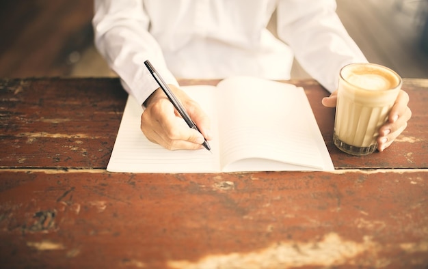 Hombre escribiendo en el bloc de notas con una taza de café en la mano izquierda en la mesa de madera