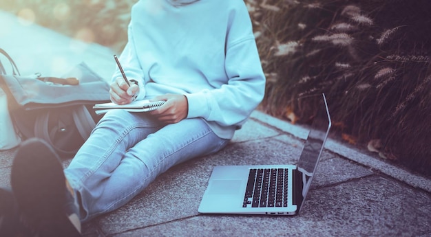 Hombre escribiendo cuaderno con computadora portátil en el parque estilo vintage Hombre hipster delgado en jeans usando un cuaderno Comunicación de autónomos Adolescente estudiante navega por Internet trabajando fuera de casa