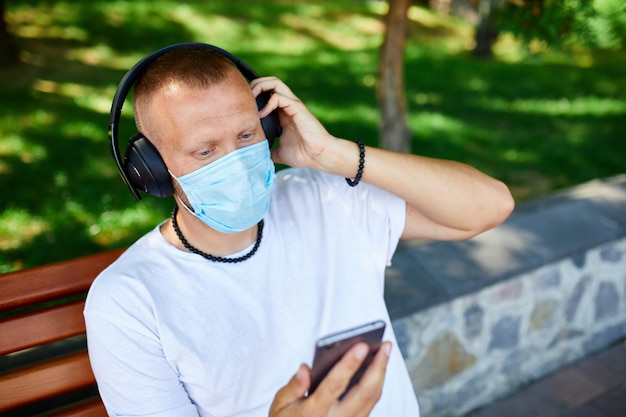 Hombre escuchando música con auriculares, usando un teléfono inteligente con máscara protectora facial al aire libre en el parque, estilo de vida nuevo normal, cuarentena, coronavirus