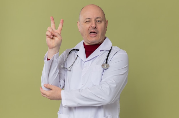 Hombre eslavo adulto alegre en uniforme médico con estetoscopio parpadeando y gesticulando el signo de la victoria