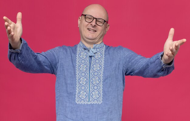 Hombre eslavo adulto sonriente en camisa azul con gafas ópticas manteniendo las manos abiertas y