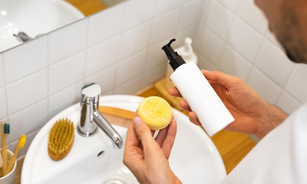 Un hombre está eligiendo entre una barra de champú sólida y una líquida. Sin plástico. Sin residuos. Ingredientes que viven con poca agua. Cuidado sostenible del cabello. Accesorios de baño. Responsabilidad por la naturaleza.