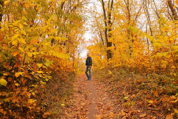 Un hombre está de pie en el camino a través del bosque otoñal, entre árboles y arbustos, una hoja de arce en sus manos, disfruta del otoño y la naturaleza