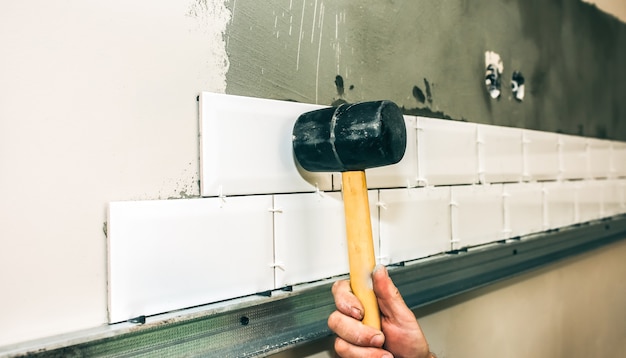 El hombre está poniendo baldosas blancas sobre el hormigón gris con la ayuda de un martillo. Mantenimiento de obras de reparación de reforma en el piso. Restauración en interior. El hombre está imprimando una superficie con un cepillo y una espátula.