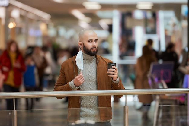 Un hombre está posando y sosteniendo una mascarilla médica que se quitó mientras toma un café en el centro comercial