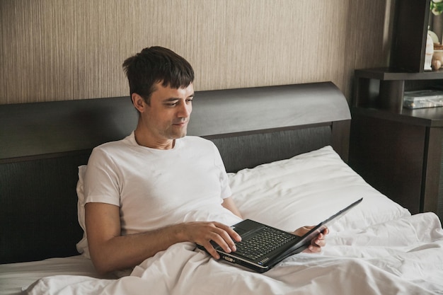El hombre está sentado en la cama en su dormitorio y trabaja en una computadora portátil